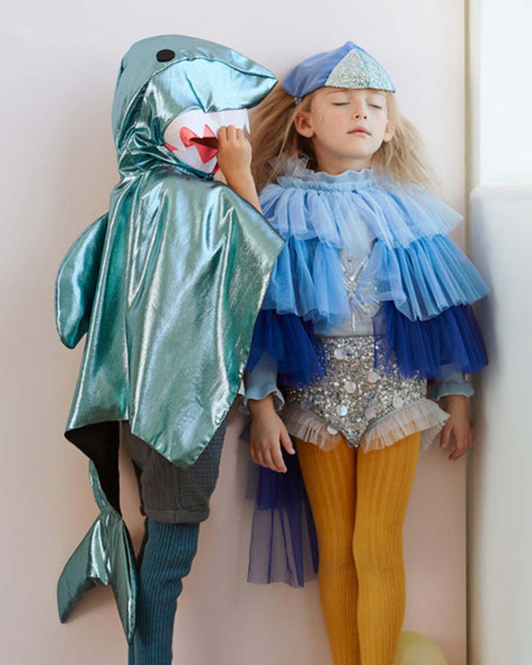 Two children in costumes: one wears the meri meri shark costume, part of a playful dress-up set, while the other sports a blue ruffled dress with a fish-scale hat and yellow tights, eyes closed, standing against a white wall.