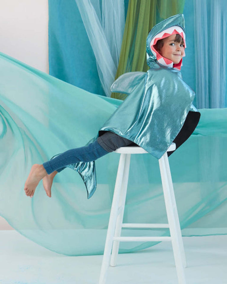 A child dressed in a shiny meri meri shark costume sits sideways on a white stool, legs extended straight out in blue tights, and is surrounded by flowing blue and green fabric.