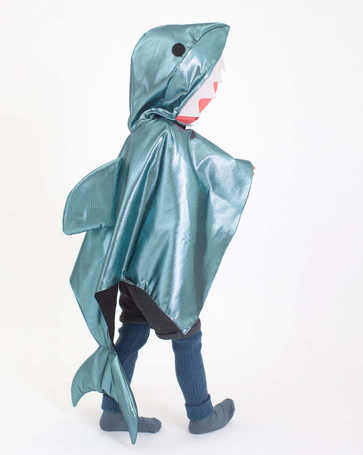 A child wears the meri meri shark costume, featuring a shiny blue design with a fin and tail, standing against a plain white background.