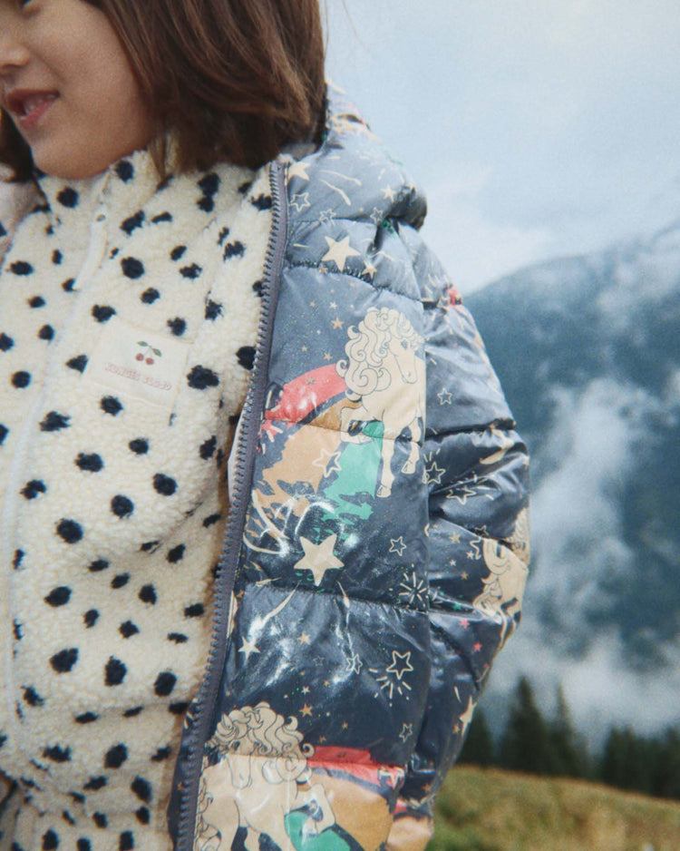 A child wearing the konges sløjd jody embroidery teddy jacket in dot lumiere—a white, OEKO-TEX certified pile fleece jacket with black spots—stands outdoors with mountains and trees in the background.