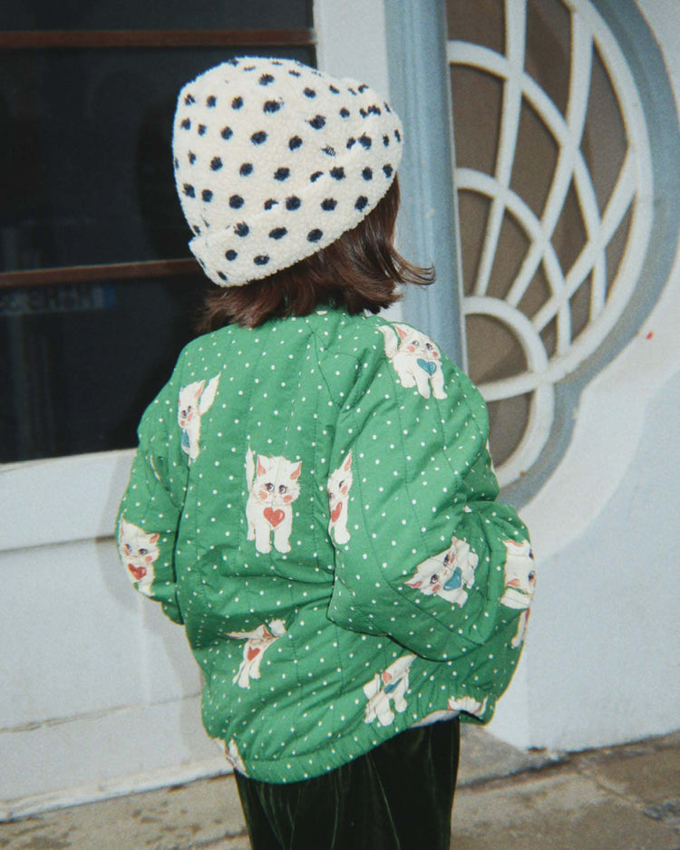 A child wearing a white hat with black dots and the konges sløjd juno printed bomber jacket in dot miao verde stands with their back to the camera in front of a decorative door.