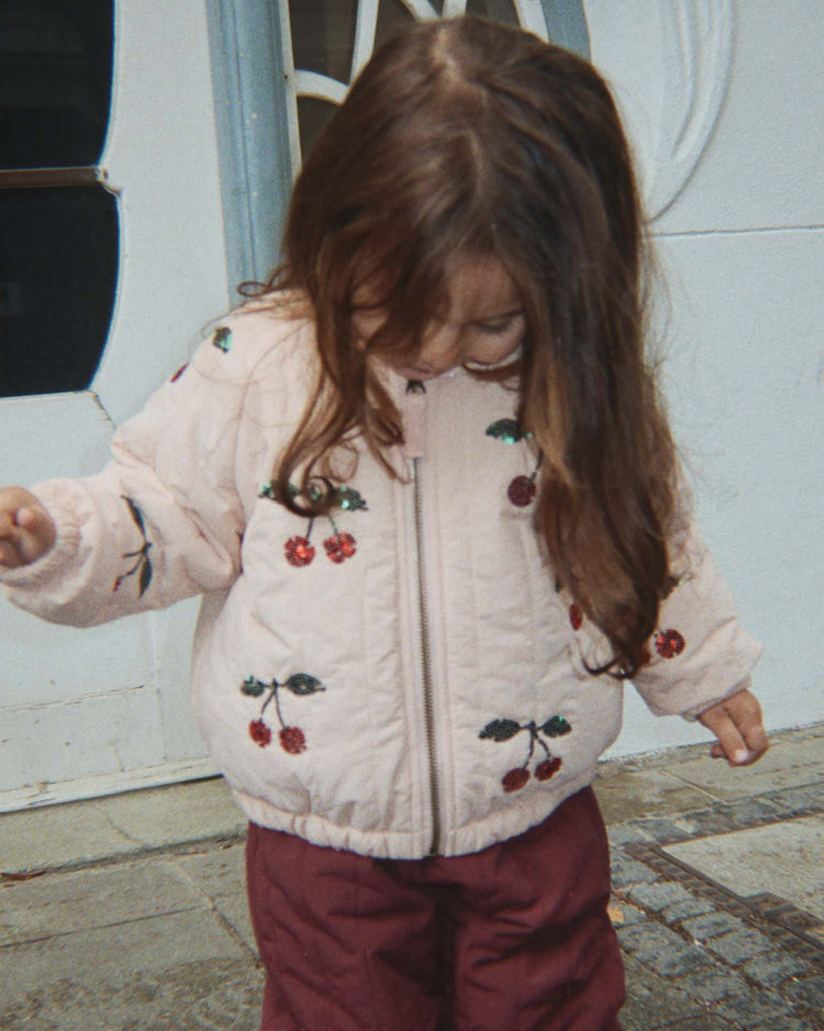 A young child with long brown hair looks down, wearing the konges sløjd juno sequin bomber jacket in cameo rose—a stylish and water-repellent outerwear piece—paired with maroon pants, standing outside on a tiled surface near a white door.