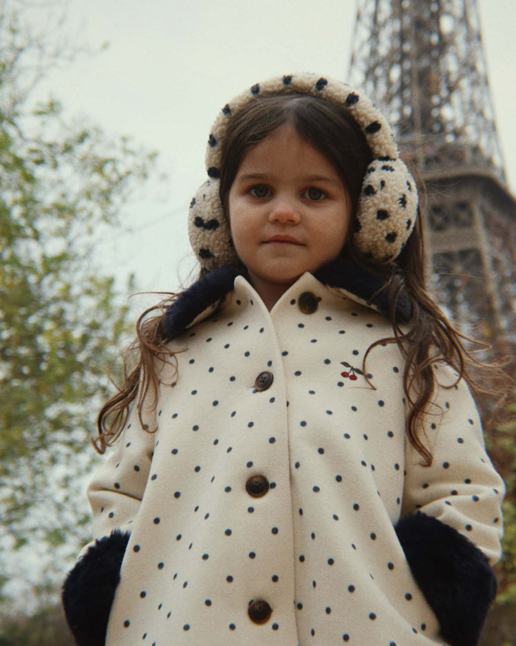 A young girl wearing konges sløjd's jody teddy earmuffs in dot lumiere stands in front of the Eiffel Tower, looking at the camera. Trees are visible in the background.