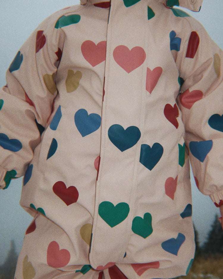 A child is dressed in the Konges Sløjd "rain snowsuit in bon coeur coloré," a light pink puffy suit adorned with colorful hearts in red, blue, green, and yellow. The snowsuit is zipped up and features a cozy fleece lining for warmth, with the child's face not shown.