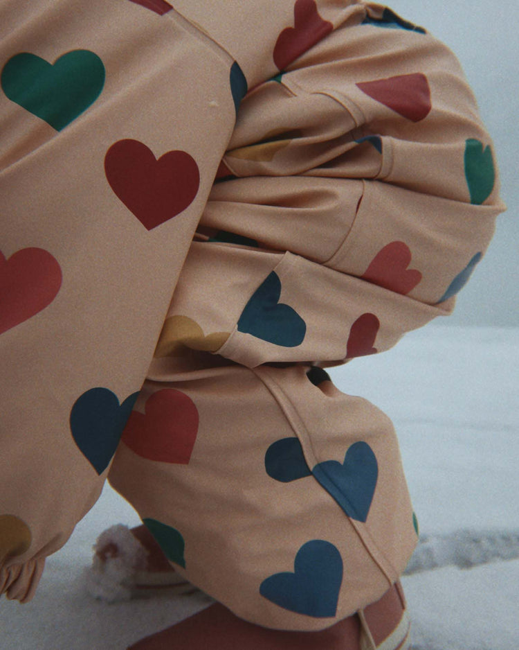A person crouches on snow, dressed in the konges sløjd rain snowsuit in bon coeur coloré, featuring beige fabric adorned with colorful heart patterns in red, blue, green, and yellow. The fleece-lined material keeps them cozy, making this snowsuit perfect for winter fun.