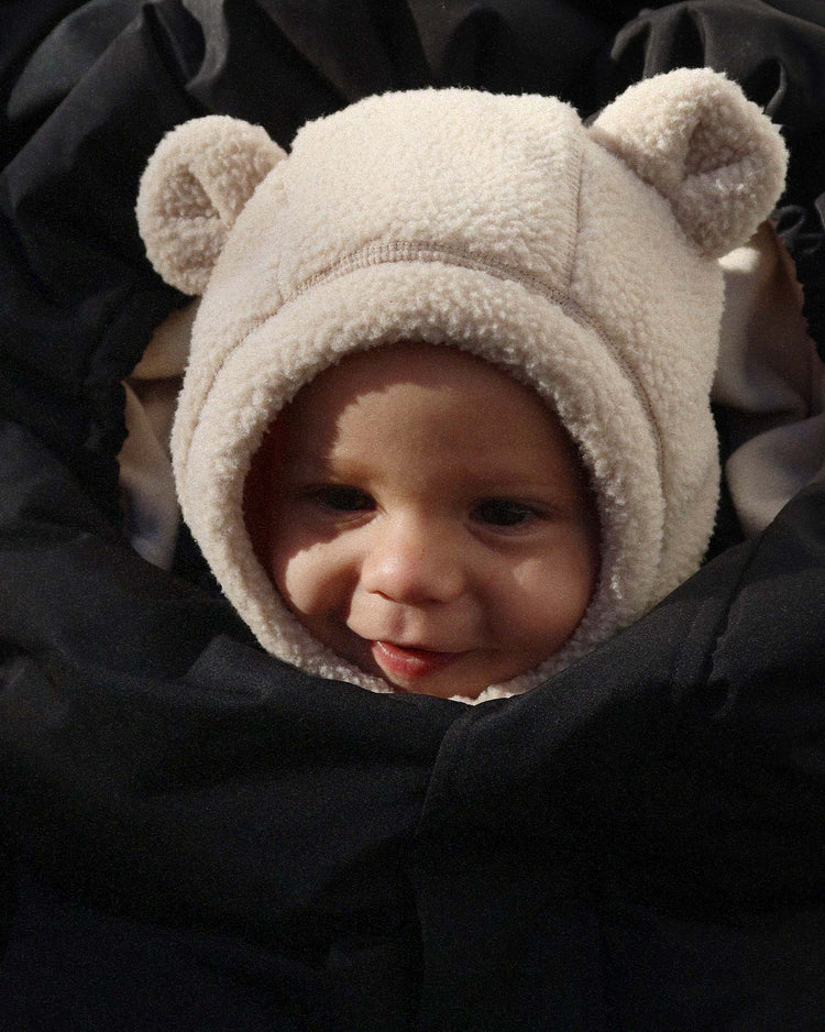 A baby wearing a fluffy safa helmet in oxford tan by konges sløjd is bundled up in a black KS105768 blanket, smiling softly.