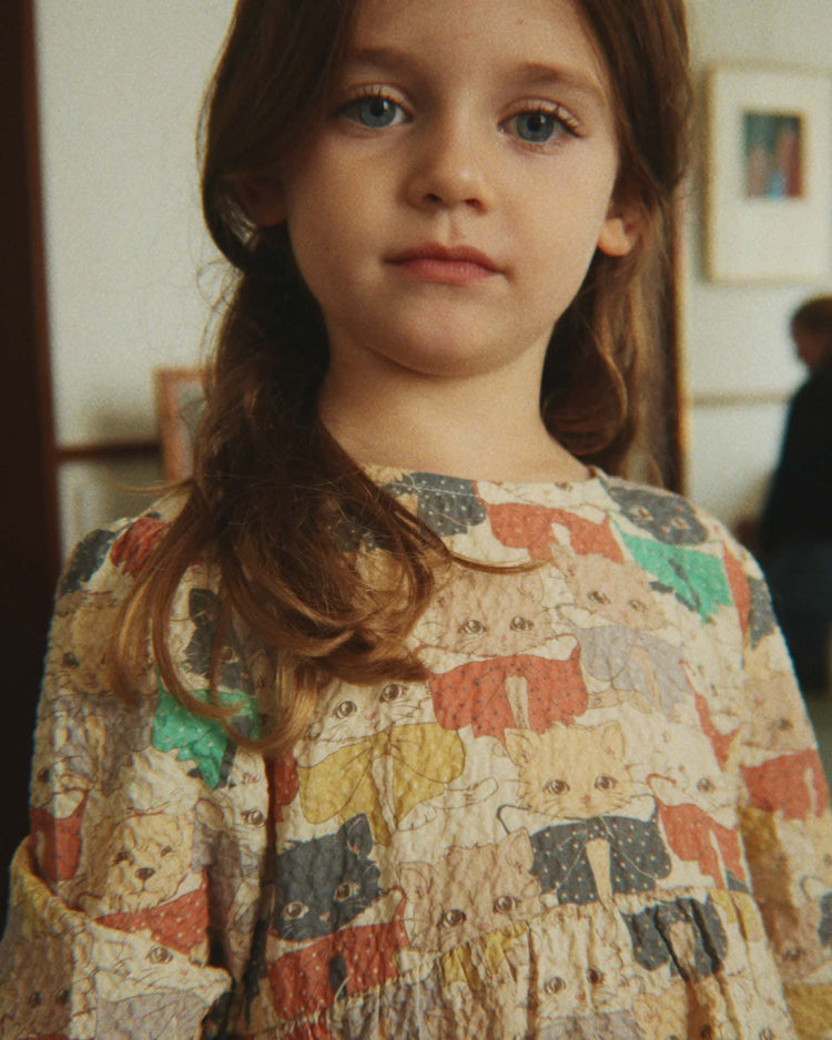 A young girl with long brown hair wears the konges sløjd elin long sleeve bow dress in "like cats and dogs," featuring a playful cat and dog pattern and voluminous sleeves, standing indoors. The softly blurred background shows framed art on the wall and another person visible in the distance.
