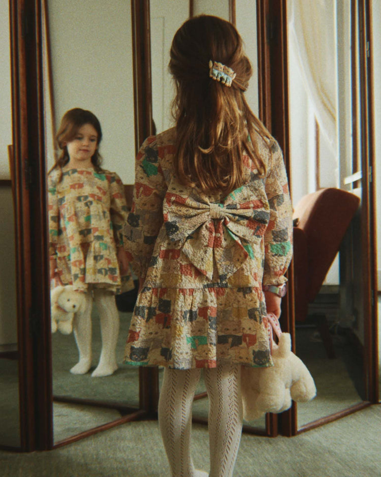 A young girl in the konges sløjd elin long sleeve bow dress in like cats and dogs, featuring voluminous sleeves and a large bow, stands before a mirror holding a stuffed toy, her reflection clearly visible.