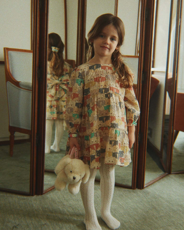 A young girl wearing the konges sløjd elin long sleeve bow dress in "like cats and dogs" with voluminous sleeves and white tights stands holding a stuffed bunny. She is positioned in front of a folding mirror, her reflection visible, within a cozy, vintage-inspired room.