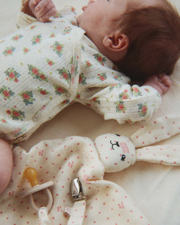 A baby in the konges sløjd minnie newborn bodysuit in rosetto, made from GOTS certified organic cotton with a floral print, lies on a soft surface next to a plush bunny toy with a pacifier attached.