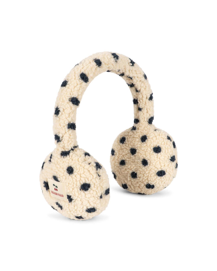 The jody teddy earmuffs in dot lumiere by konges sløjd feature a cream-colored, fluffy textured fabric adorned with black polka dots. Designed for comfort, these earmuffs include an adjustable headband and are finished with a small tag bearing red text on one side.
