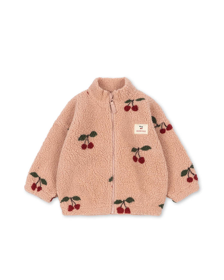 The konges sløjd Jody Teddy Jacket in Ma Grande Cerise Blush is a beige, fluffy pile fleece jacket for kids, featuring an all-over red cherry and green leaf print and a high collar. Made from recycled polyester, it includes a small rectangular patch sewn on the chest. The jacket is shown laid flat on a white background.
