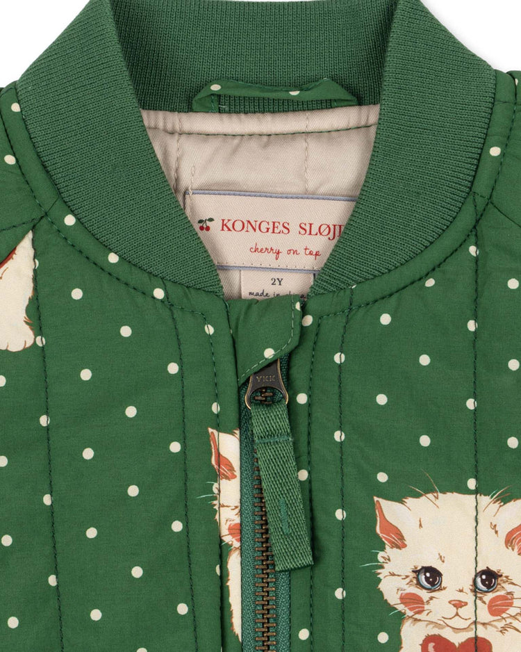 Close-up of the juno printed bomber jacket in dot miao verde by konges sløjd, featuring green quilted fabric with white polka dots, a zipper, and an adorable kitten print. This lightweight thermo jacket is both wind-resistant and water-repellent for comfort in any weather. Label reads: konges sløjd, cherry on top, 2Y.