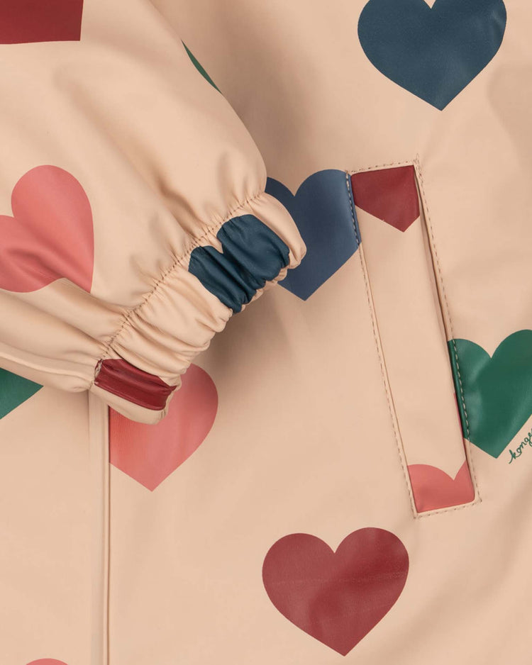 A close-up of the konges sløjd rain snowsuit in bon coeur coloré shows its beige fabric adorned with vibrant blue, red, and pink heart prints. The image highlights details such as an elastic cuff and a side pocket seamlessly blending into the playful heart pattern, while showcasing the cozy fleece-lined interior designed for extra warmth.