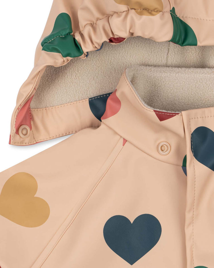 Close-up of the "rainy palme set girl in bon coeur coloré" by konges sløjd, featuring a beige raincoat adorned with colorful heart patterns. The hood and collar are lined with soft white fleece, offering added warmth and comfort to this stylish, wind-resistant waterproof set.