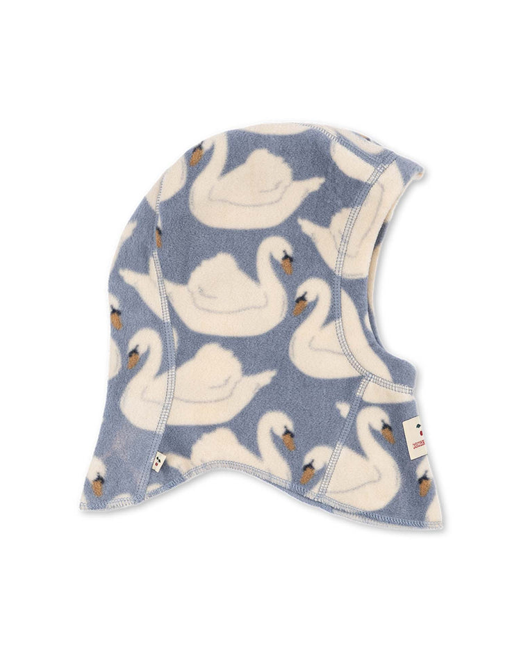 The konges sløjd tavi fleece balaclava in swan is a blue fleece bonnet featuring a pattern of white swans with orange beaks. The balaclava has a rounded, snug shape and visible stitching along the edges.