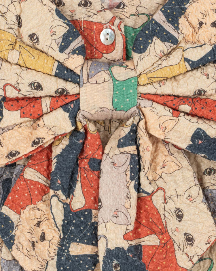 A close-up of fabric from the konges sløjd elin long sleeve bow dress in like cats and dogs, showcasing a playful pattern of cats and dogs in polka dot outfits, highlighted by a button and a large central bow—evoking the look of an organic cotton dress with voluminous sleeves.