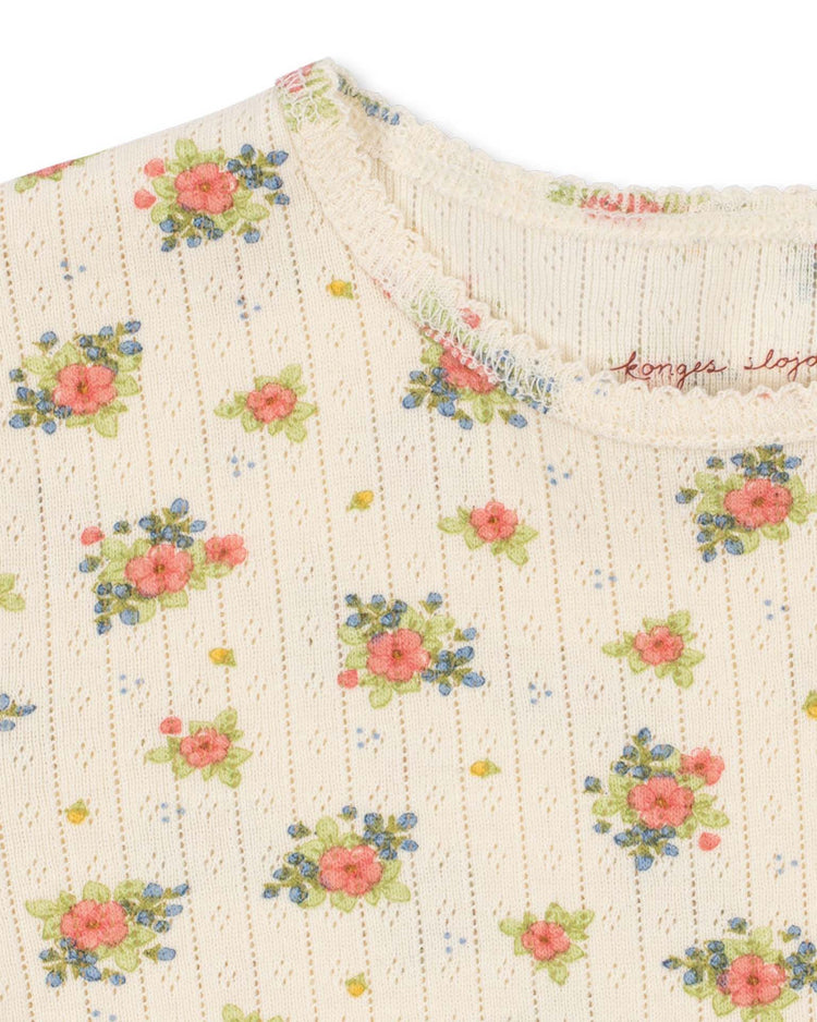 Close-up of the minnie blouse in rosetto by konges sløjd, featuring cream-colored organic cotton with a ribbed texture, small floral clusters in pink, blue, and green, and scalloped lace trim around the neckline.