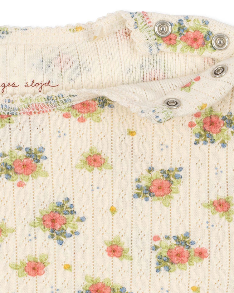 Close-up of the minnie bodysuit in rosetto by konges sløjd, crafted from cream-colored ribbed organic cotton adorned with delicate pink, blue, and yellow floral prints. The fabric features metal snap buttons and a partially visible stitched label with red text—perfect for baby essentials made from GOTS certified materials.