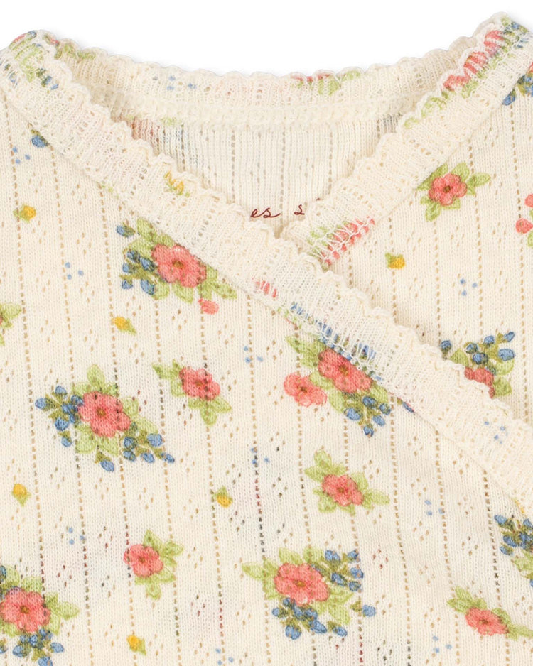 Close-up of the cream-colored, ribbed organic cotton fabric of the "minnie newborn bodysuit in rosetto" by Konges Sløjd, featuring a delicate floral pattern in pink, blue, and green. This baby bodysuit offers scalloped trim along the edges and a chic crossover design detail, crafted from GOTS-certified materials for exceptional softness and reassurance.