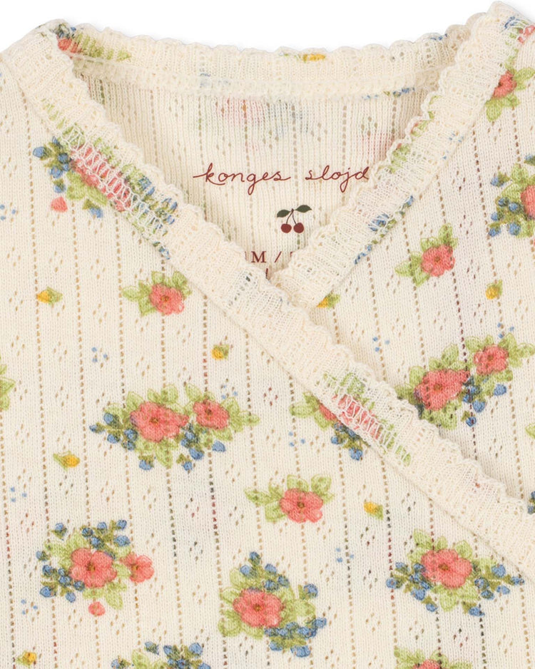 Close-up of the minnie newborn onesie in rosetto by konges sløjd—a cream-colored, ribbed wrap-style onesie with lace trim. Crafted from GOTS certified organic cotton, it features a delicate floral print with pink, blue, and green flowers. The konges sløjd logo is printed on the inside.