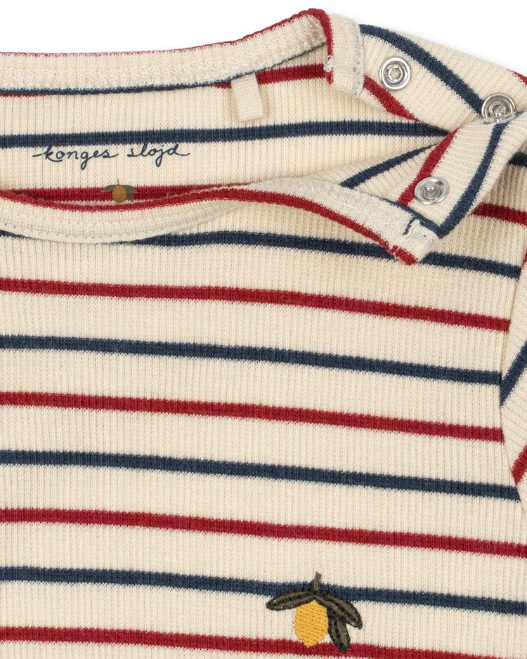Close-up of the voco modal bodysuit in tricolore bleu by konges sløjd—a cream-colored piece with red and navy horizontal stripes, metal snaps on the shoulder, and a small yellow lemon embroidery near the bottom. Crafted from a soft modal blend, it features an inner label with the konges sløjd name.