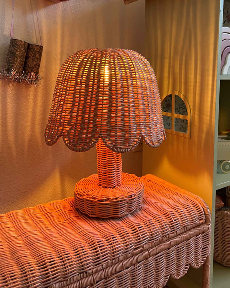 The dinkum dolls petal lamp in seashell pink features a scalloped shade and emits a warm glow, creating cozy nursery lighting when placed on a table against a beige wall.