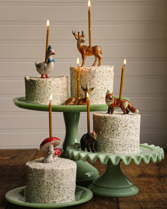 Three tiered cakes with animal figurines as toppers and lit candles on rustic green stands.