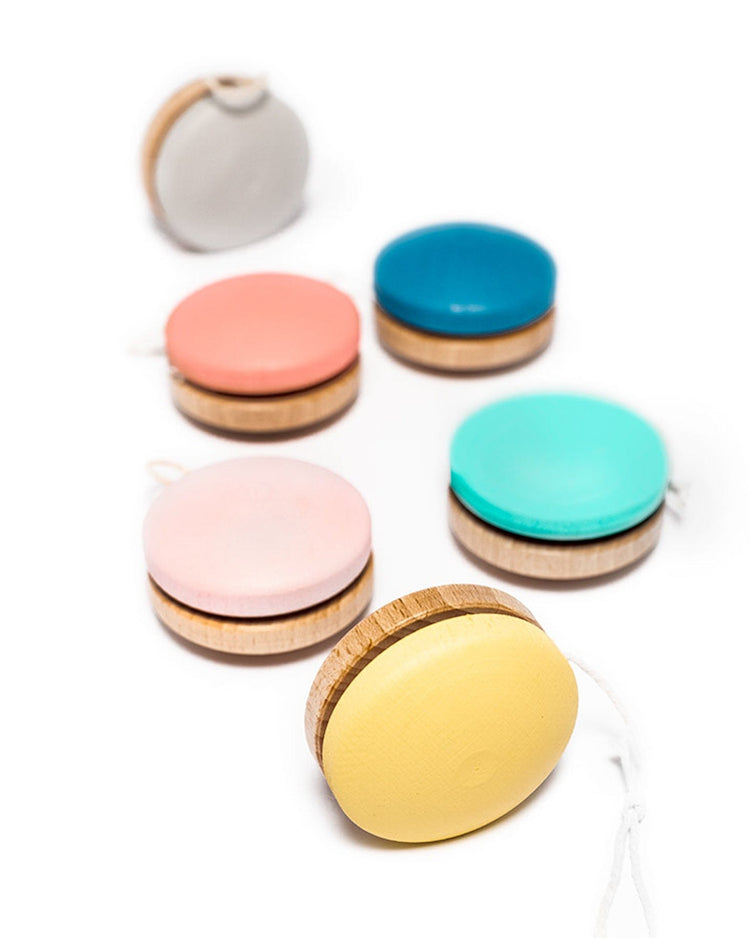 A group of me & mine wooden yo-yos, painted in vibrant colors such as gray, pink, blue, turquoise, light pink, and yellow using eco-friendly paint, are neatly arranged on a white background. These sustainable toys include the popular yo yo in turquoise among other hues.