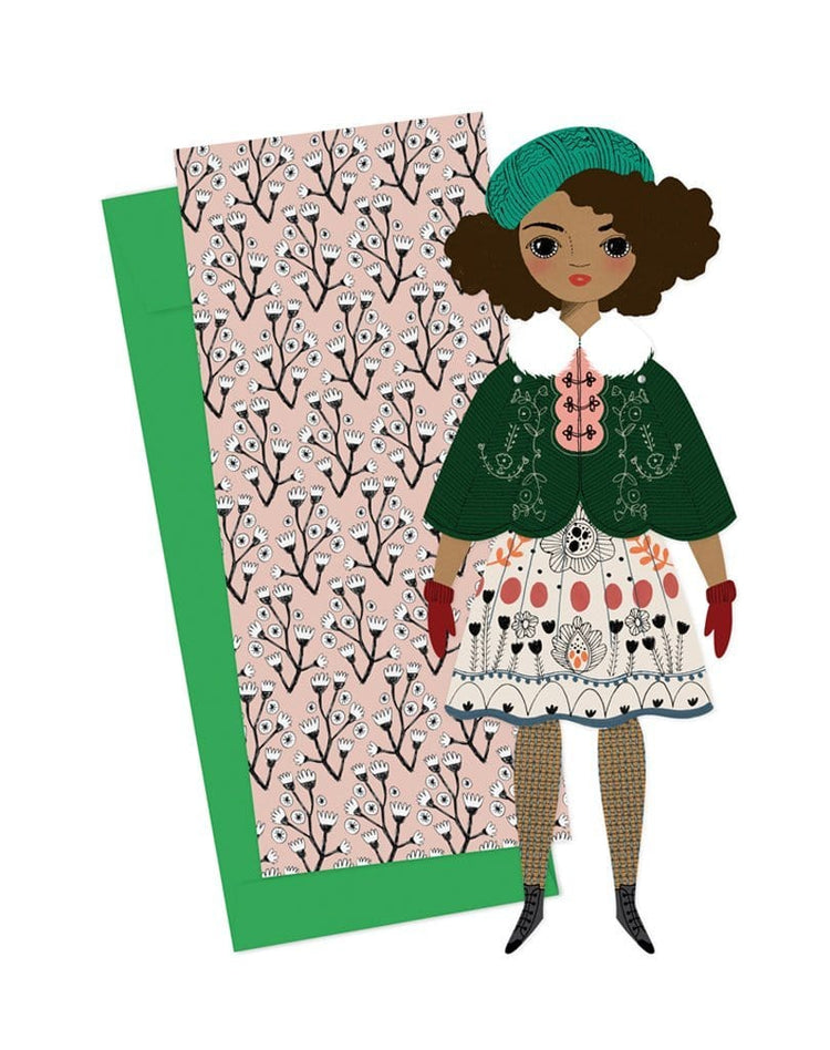 Illustration of a handmade Of Unusual Kind Noelle Mailable Paper Doll with a patterned background, wearing a green hat, green jacket, and a dress with floral motifs.