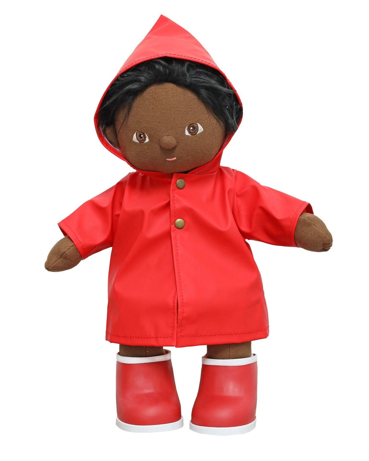 A soft doll with brown skin, black hair, and brown eyes stands facing forward, dressed in the dinkum dolls dinkum doll ahoy raincoat in red—waterproof and made for 14-inch dolls—with matching red rain boots.