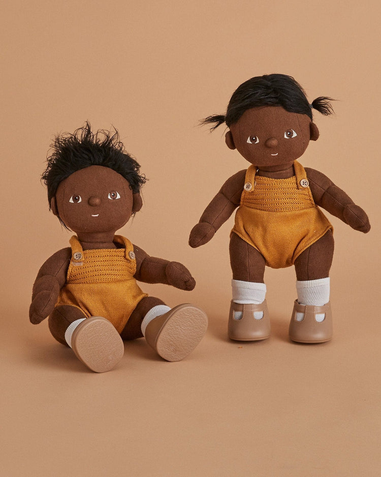 Two dark-skinned dinkum dolls - tiny by dinkum dolls with black hair, yellow overalls, and white socks. One posable doll has short tousled hair and sits; the other stands upright in shoes with pigtails. Beige background.
