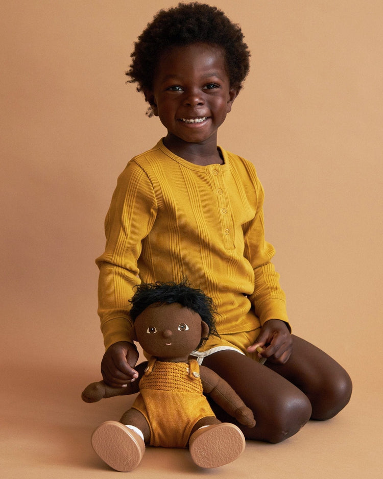 A young child in a mustard yellow long-sleeve shirt sits smiling on the floor, holding a dinkum dolls dinkum doll - tiny dressed in yellow overalls against a warm beige background.