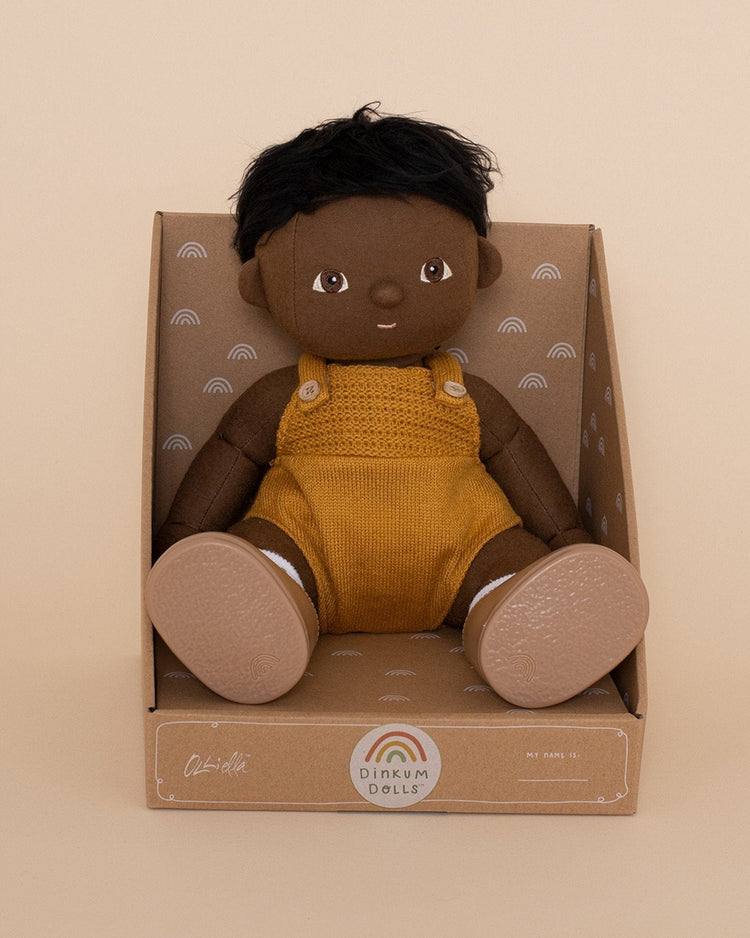 A soft, posable dinkum dolls "dinkum doll - tiny" with brown skin and short black hair in mustard overalls sits upright in a rainbow-patterned box labeled on the front.