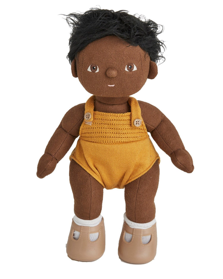 A dinkum dolls dinkum doll - tiny with dark brown skin, short black hair, dressed in a yellow knitted romper with buttons and light brown shoes, stands upright and faces forward.