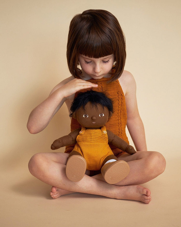 A young child with short brown hair, dressed in orange, sits cross-legged on a beige floor and gently touches the head of a dinkum dolls dinkum doll - tiny, which is wearing yellow overalls.