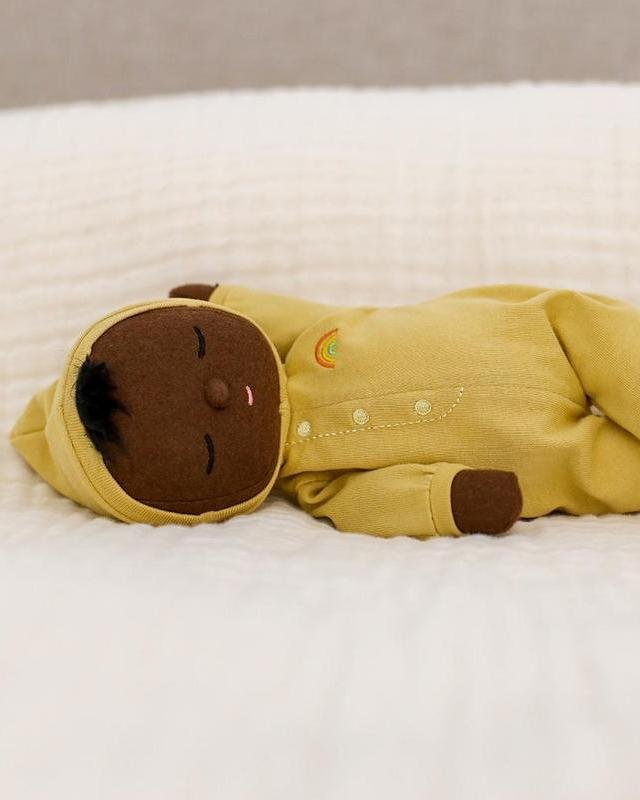 The dinkum dolls dozy dinkum mini is a soft brown doll with black hair, pink mouth, and closed eyes, dressed in a yellow hooded onesie with a rainbow embroidery—a perfect newborn baby gift.