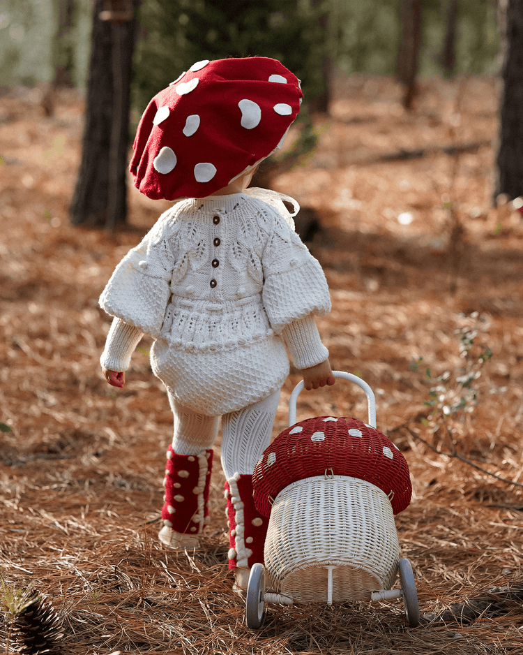 A small child in a red mushroom hat and boots strolls the forest, pulling the dinkum dolls rattan mushroom luggy in red—a whimsical, wheeled storage basket perfect for toys on any woodland adventure.