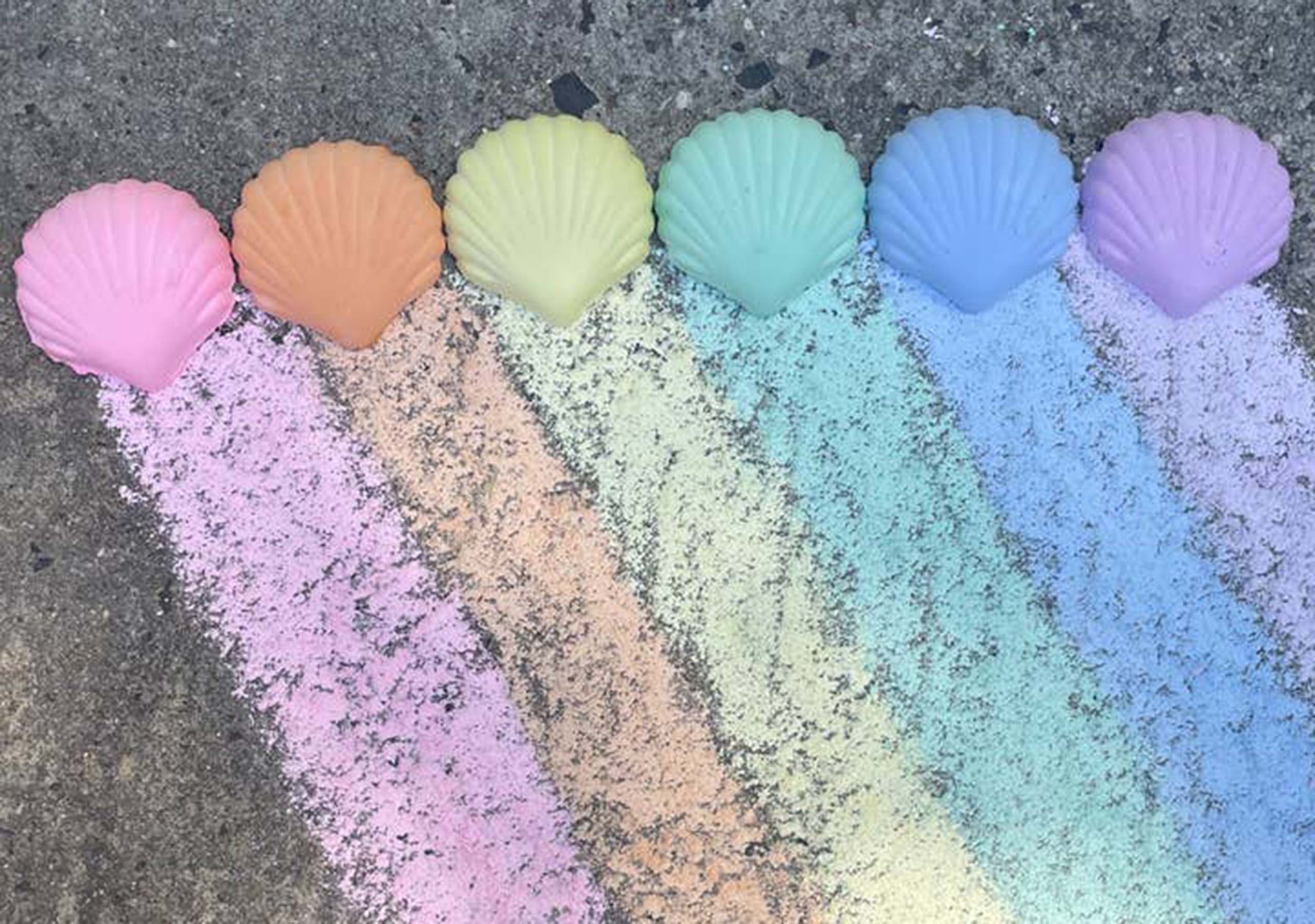Six pastel-colored seashell molds arranged in a row on pavement, each with matching chalk powder creating a rainbow pattern underneath them: pink, orange, yellow, green, blue, and purple.