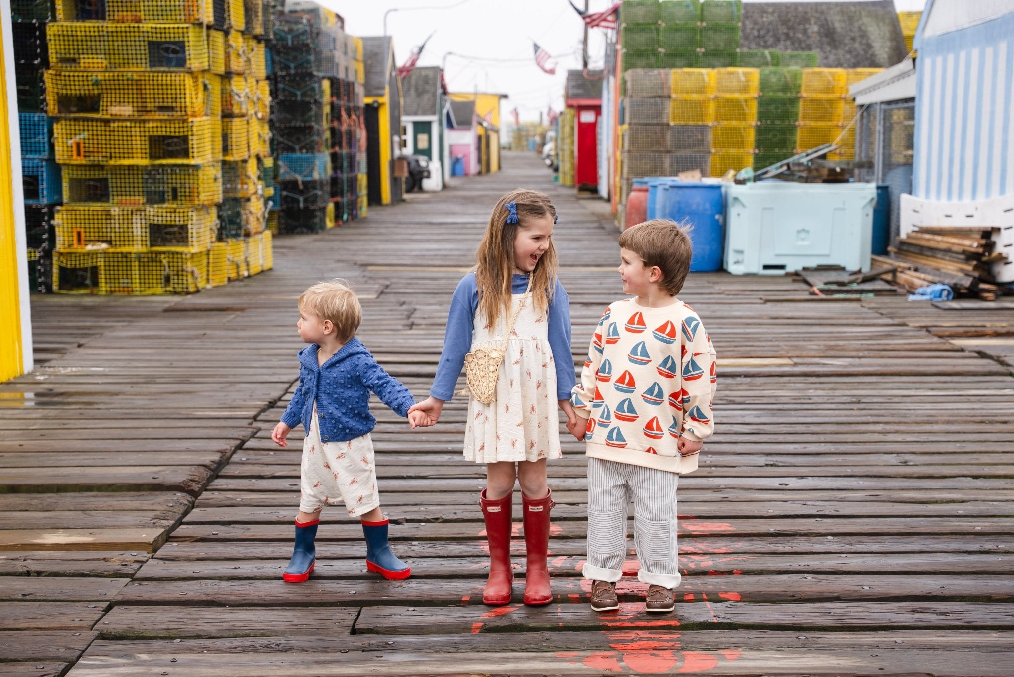 Three young children stand on a wooden dock, holding hands and smiling. Colorful lobster traps and fishing gear are stacked along both sides. The kids wear rain boots and patterned clothes.