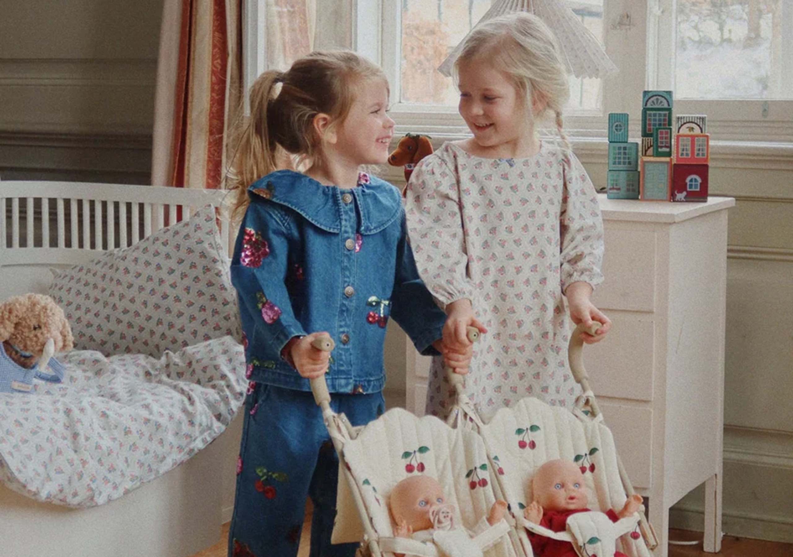Two young girls smile at each other while holding toy strollers with baby dolls. They stand in a cozy, sunlit room with a bed, dresser, and toy buildings by a window in the background.