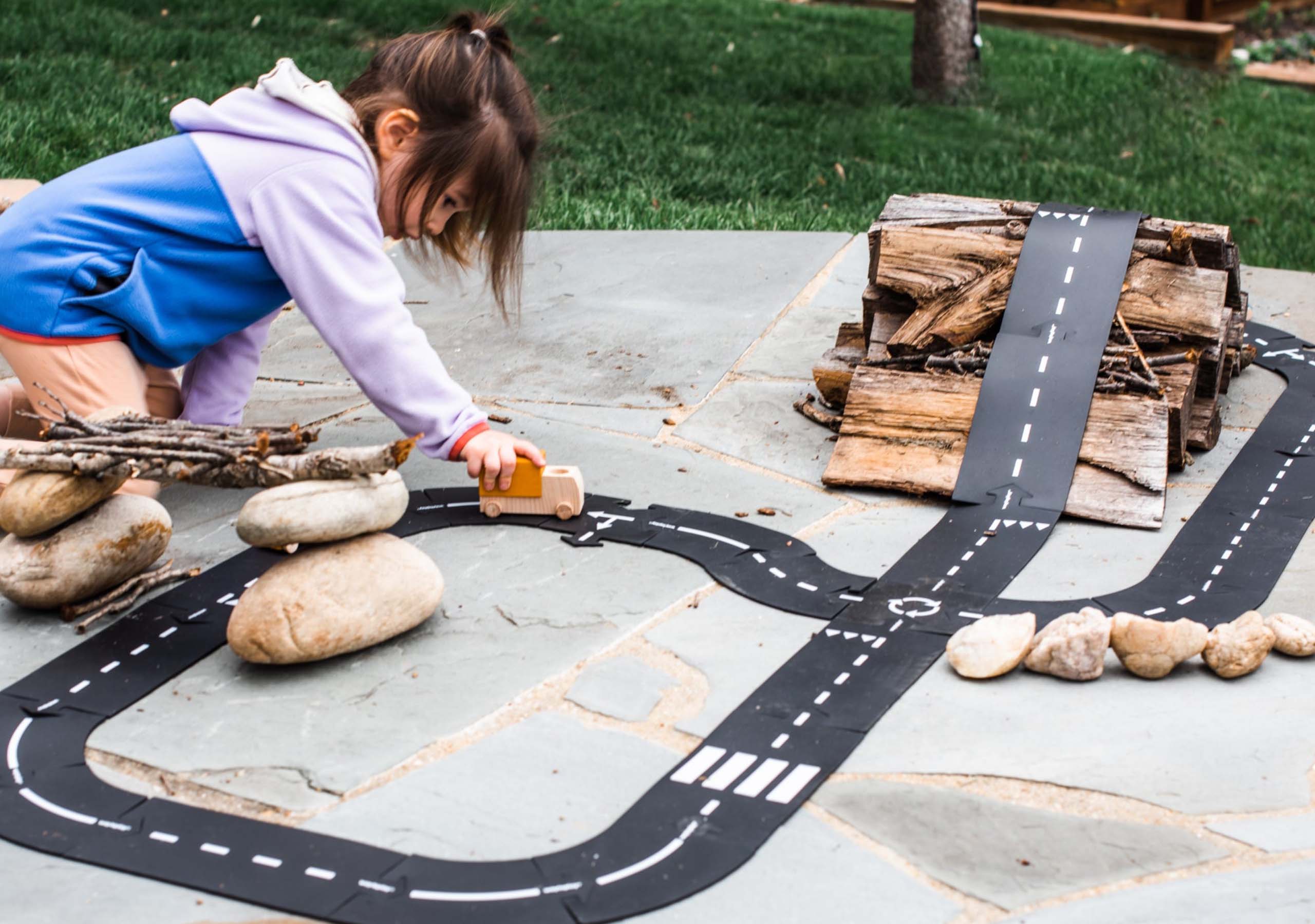 A young child stands by a large window, placing a toy car on a black track attached to the glass. Colorful curved toys and a potted plant are nearby, with a garden visible outside.