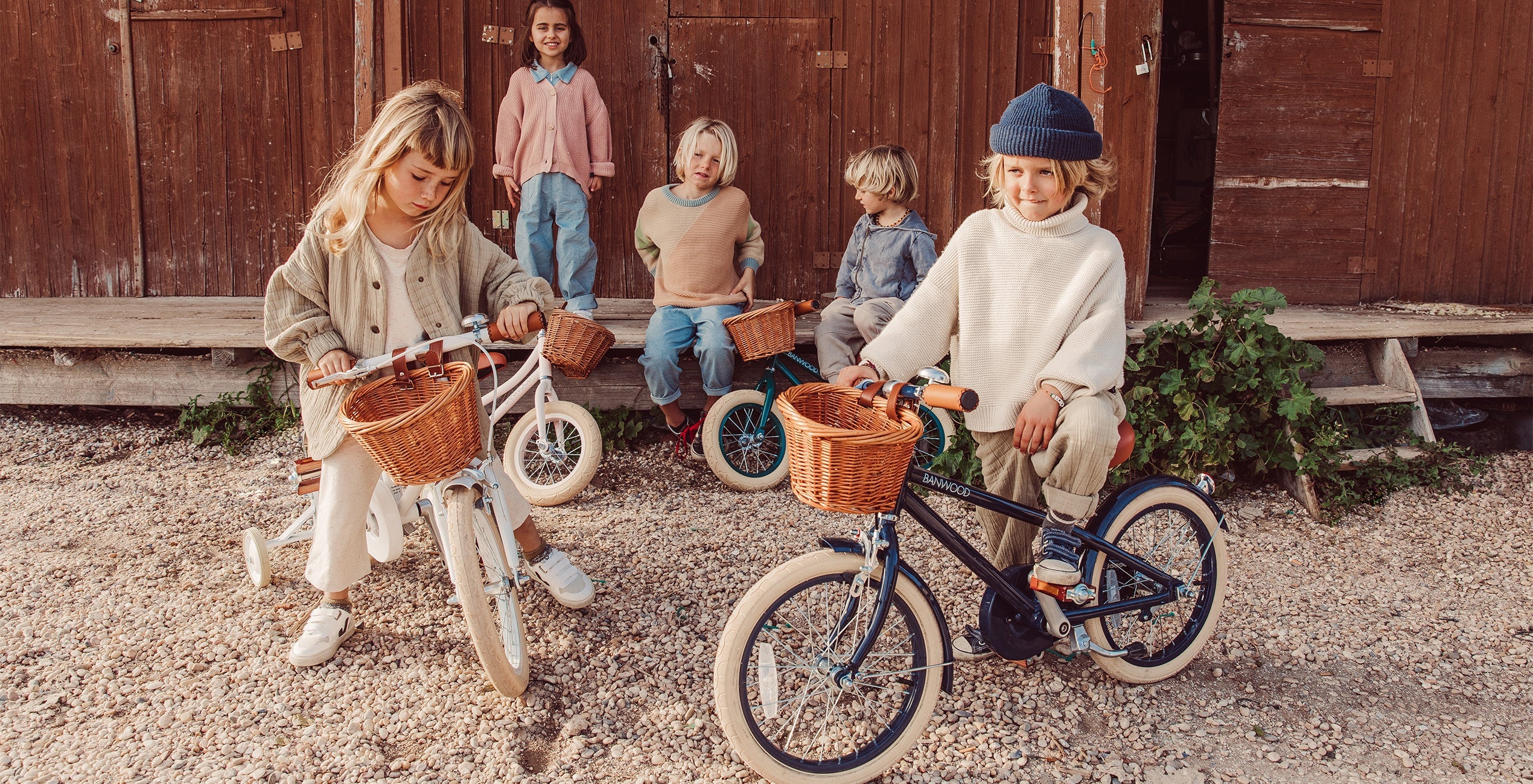 Five children are gathered outside a wooden building. Two are riding bicycles with wicker baskets, while three are sitting on the steps in the background.