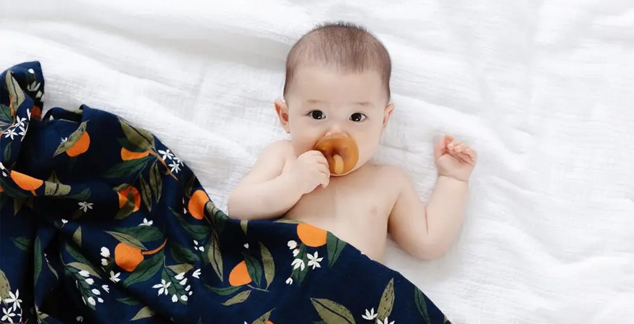 A baby lies on a white surface, partially wrapped in a navy blue blanket with an orange and green leaf pattern. The baby is sucking on a pacifier and looking upwards.