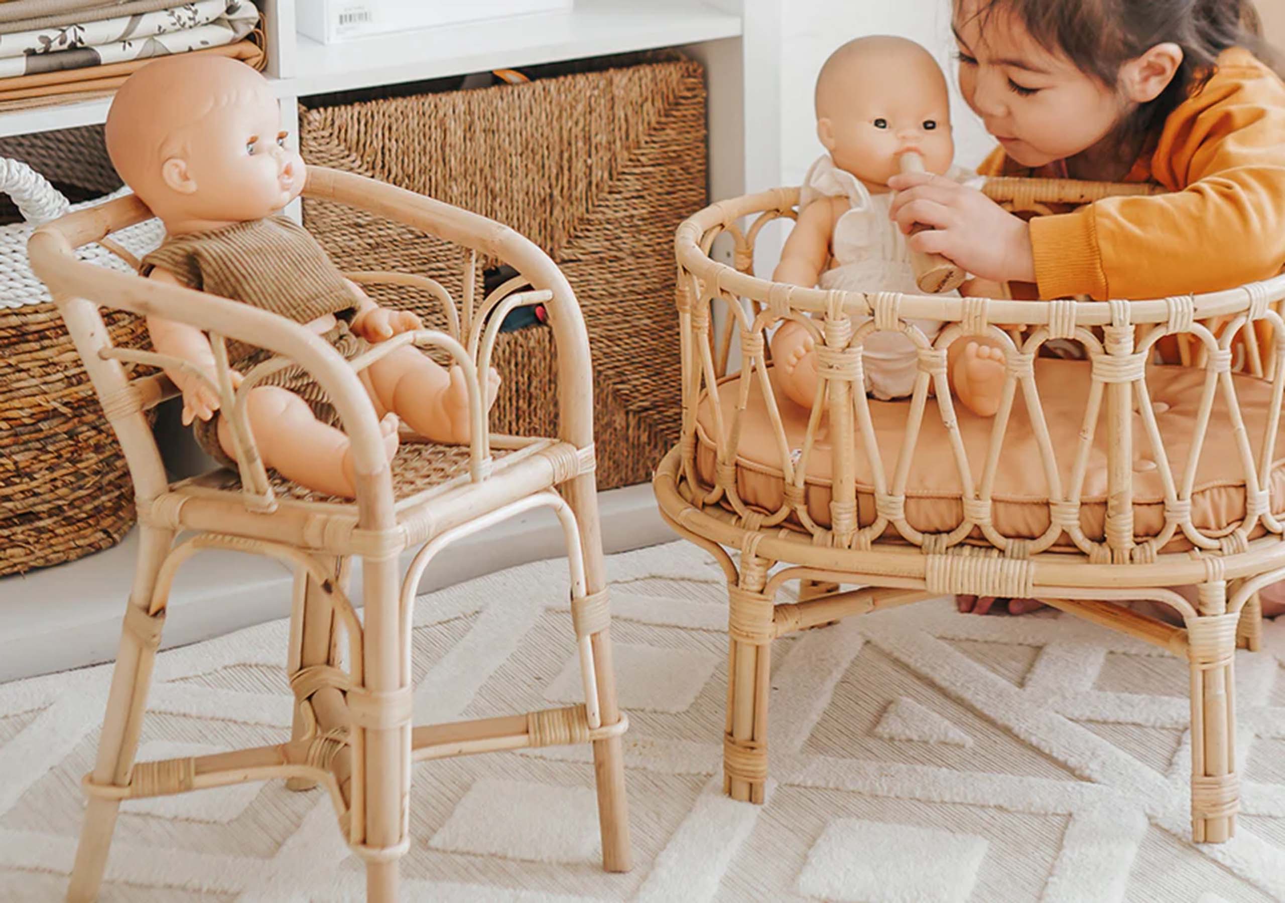 A young child plays with a doll in a rattan crib while another doll sits in a matching rattan high chair. Woven baskets and a shelf are visible in the background.