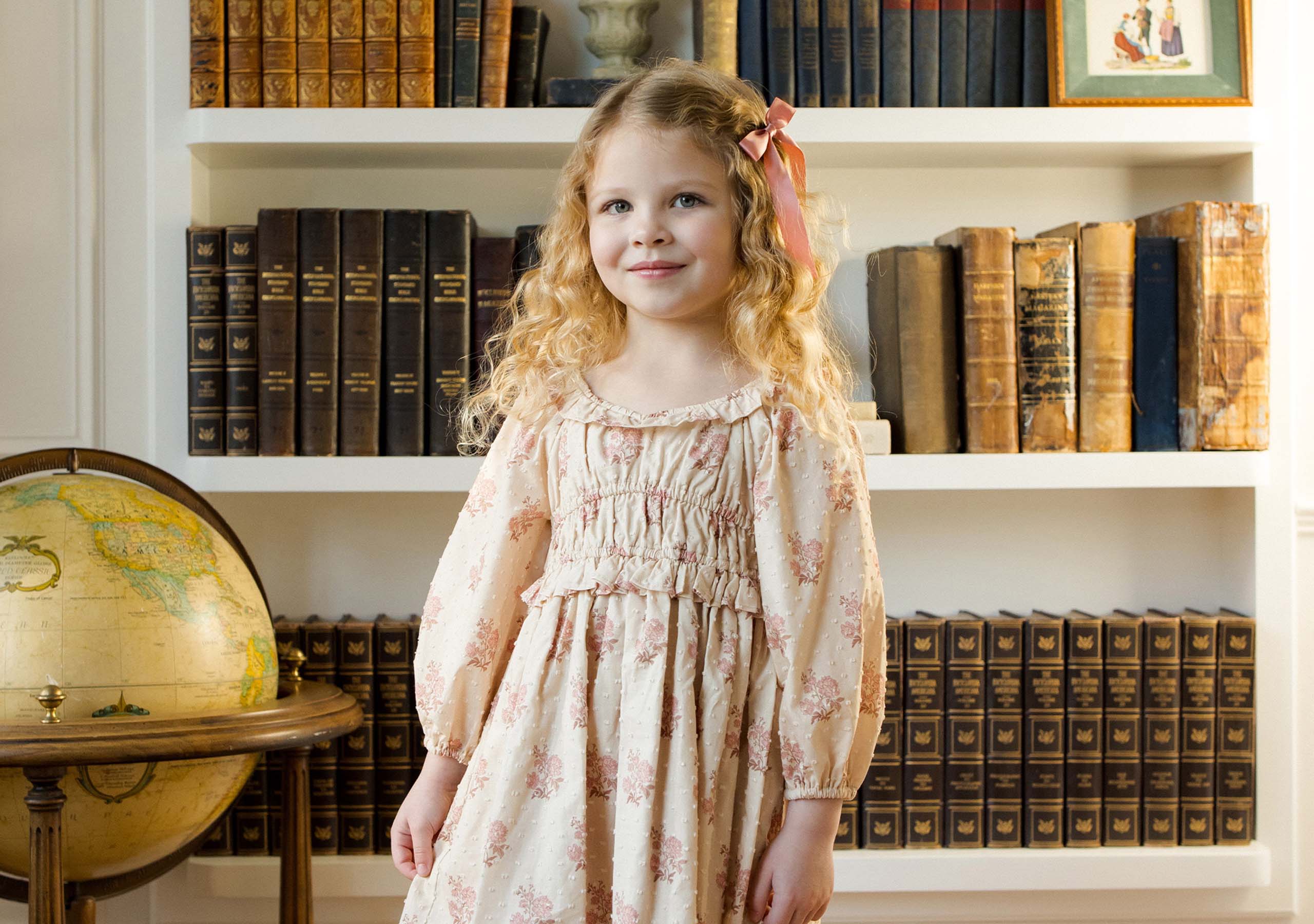A young girl with curly blonde hair, wearing a light pink floral dress, stands smiling in front of bookshelves filled with old books and a globe.