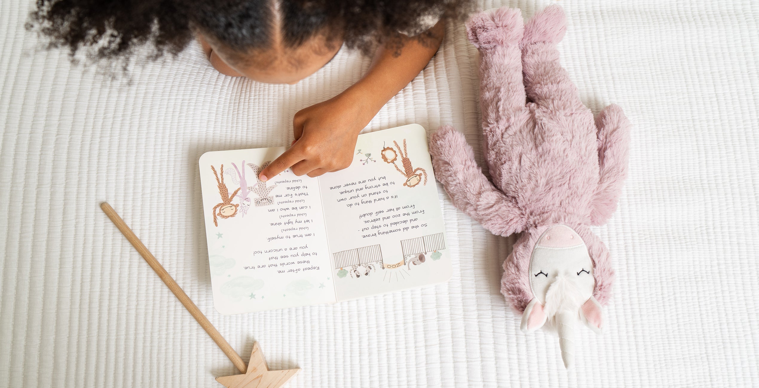 A child lies on a white surface reading a storybook, with a stuffed unicorn toy and a wooden wand placed beside them.