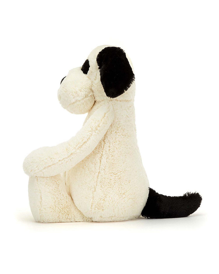 The Jellycat Bashful Rufferty Black + Cream Puppy Really Big is a soft, plush stuffed dog with cream fur, black floppy ears, a black tail, and a black spot over one eye, shown sitting upright against a white background.