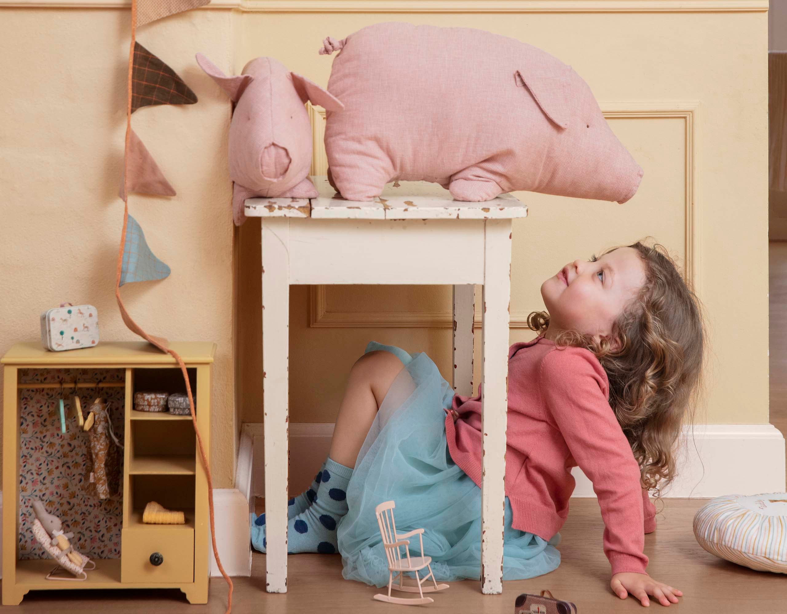 A child lies beneath a small table, gazing up at a large plush pig atop it in a cluttered playroom.