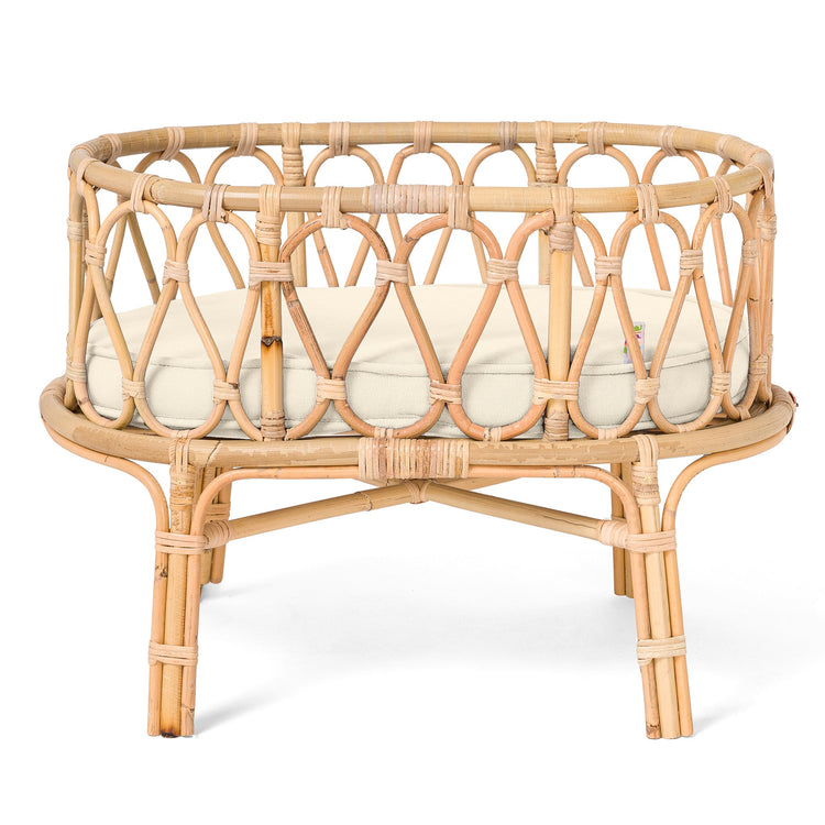 Round rattan pet bed with an open-weave design, elevated legs, and a thick cream-colored cushion creates a cozy spot for your furry friend—perfect for the playroom or next to the Poppie Crib Classic Collection by poppie toys, nestled among their favorite soft toys.